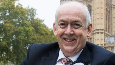 A close up image of Sir Wayne David in front of the UK Parliament. He is smiling and wearing a dark suit, a white shirt and a red patterned tie. 