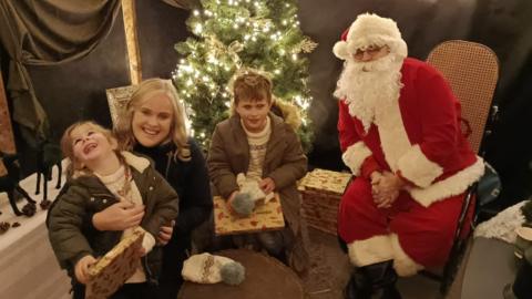 Two children holding presents, pictured with their mother and Santa Claus in a grotto man dressed as Santa Claus