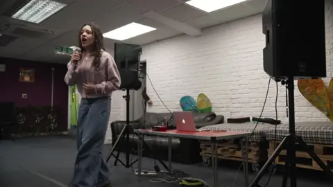 A young girl with long brown curly hair, a pink hoodie and blue wide-legged jeans sings into a microphone, with a laptop, speakers and music equipment in the background