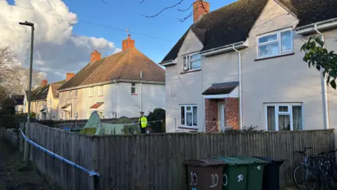 A police officer stands next to a white house, which is surrounded by police tape.