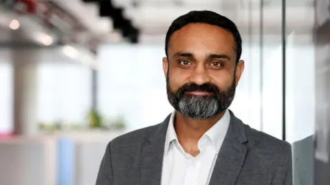 Cancer Research UK Turabi has short black hair and black moustache and beard with some grey hairs. He is wearing a grey blazer and a white shirt which is unbuttoned.