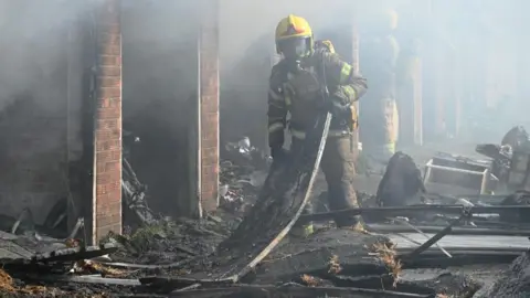 Norfolk Fire and Rescue Service A firefighter in full breathing apparatus and a yellow helmet holds a piece of metal sheeting, which is in the middle of debris outside a row of burned-out garages. Another firefighter is standing in the entrance to another garage just behind him. There is a haze of smoke in the air around them.