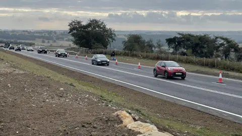 Vehicles travelling on a newly laid road with red and white cones lining the road on the far right of the picture. To the left of the picture is soil and a few light brown stones with short green grass.