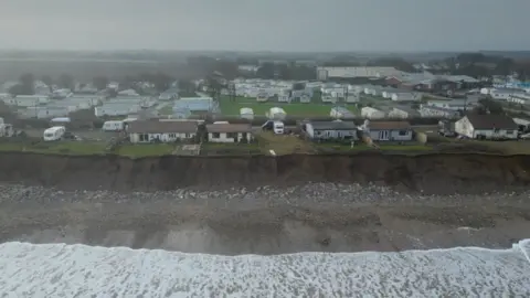 Holiday chalets and caravans are close to the edge of a cliff-top. Below them is a sandy beach with white stones and the tide coming in, which is foamy and white.