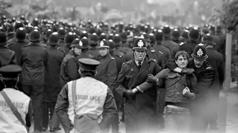 PA A black and white photograph from Orgreave when violence broke out. A sea of police helmets, adorned with constabulary badges, stretches out into the distance. In the foreground, two officers - dressed in trench coats - escort away a young man - dressed in a denim jacket and jeans. One officer wraps an arm around his neck, whereas the other uses both hands to restrain one of his arms. Two more men are stood to the side of the scene, wearing vests that read: "Ambulance."