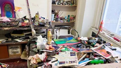 A room in Glenys' house where she paints with a desk and chair. Every surface is covered in paint, paper and painting tools