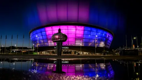Glasgow 2026 The OVO Hydro arena in Glasgow is lit in purple and blue at night, reflected in a puddle in the foreground, with a large sculpture standing outside the building.