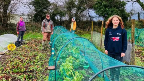 Steve and his family are seen weeding a vegetable patch with small shovels. Lola, 13, is at the front wearing a blue jumper, her dad Steve is behind her while opposite them is Lola, 16, and mum Rebecca at the back. Around them nets are covering vegetables and a black bucket stands to the left. 