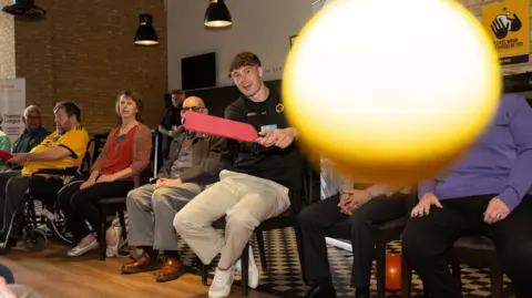 A man wearing a black Wolverhampton Wanders shirt is holding a red cricket bat while sat down next to elderly people in a room.