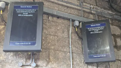 A grey wall with two electronic signs. They both say " Special notice, no service in Cornwall until at least 16:00 following Storm Goretti." 