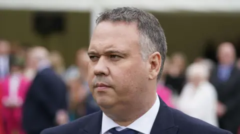 A man with short grey and black hair looks off to the side of the camera. He is wearing a dark-coloured suit jacket, a white shirt, and a tie. The background is out of focus, but features a number of people standing. 