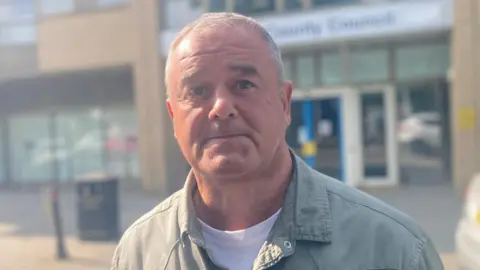 BBC/Stuart Whincup David Moore outside the court. He looks full of emotion. He is wearing a khaki green shirt and white t-shirt and has short greying hair.