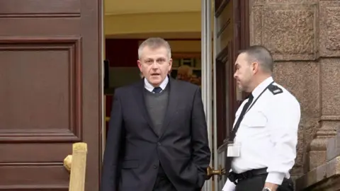 BBC Deputy Philip Ozouf is walking out of a wooden door at Jersey's Royal Court building. He is wearing a dark suit and grey jumper and looking into the camera with his mouth open. A court usher in a black and white uniform is standing by the door.