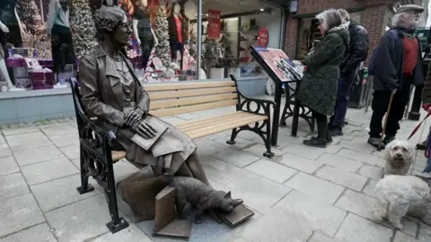 The bronze statue sitting on the bench in the pedestrianised shopping street. The figure has her legs stretched out and resting on a pile of books with a cat, also part of the sculpture, rubbing its head on her feet. To the right of the picture, two people are reading an information board.