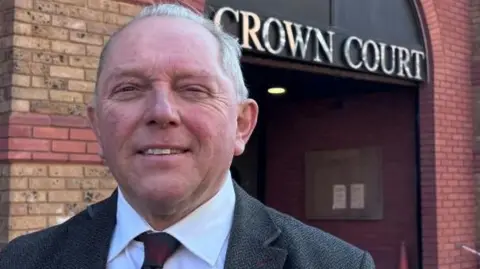 Derek Bennett: a grey haired man wearing a grey jacket and red tie outside a red-and-brown brick building. The words "crown court" are written above the entrance to the building in capital letters. 