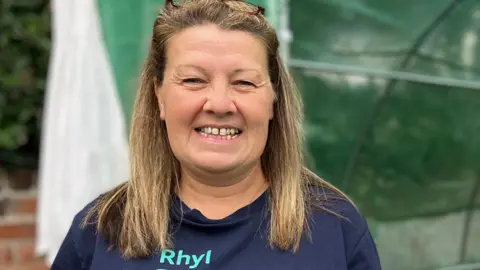 Penny standing in front of a polytunnel with green netting. She is smiling at the camera. She has a navy blue T shirt and shoulder length light brown hair.