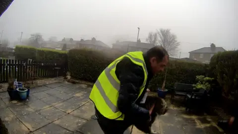 CCTV footage of a man in a high-vis vest picking up a cat in a garden