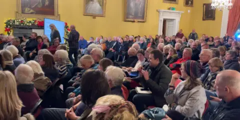 Grand room with yellow wallpaper and ornate paintings, rows of people sit and face the left of the image like they are watching a presentation