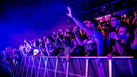 A crowd behind a barrier watches a gig at the Roundhouse in London. They are holding there arms in the air, filming on phones and bathed in blue lighting.