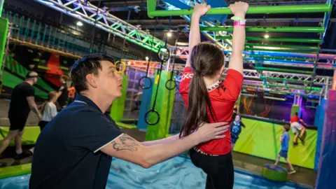 Man helps a child onto monkey bars at assault course.