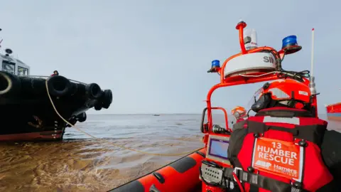 Humber Rescue A Humber Rescue lifeboat can be seen on the right hand side out at sea with the back of one man men operating it and wearing a red lifejacket and helmet. A large vessel can be seen in the distance on the left with rope attached to it from the lifeboat.