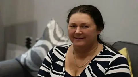 A woman with brown hair sits on a grey sofa. She wears a black and white striped top. Her hair is tied back into a ponytail.