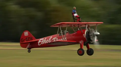 A red biplane taking off from a runway with a grey haired woman seated on a platform above the upper wing.