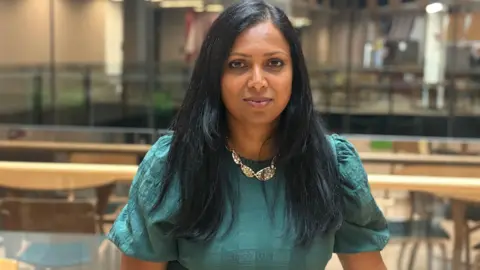 A woman is looking into the camera. She's wearing a green dress and a siler necklace. She has long black hair.