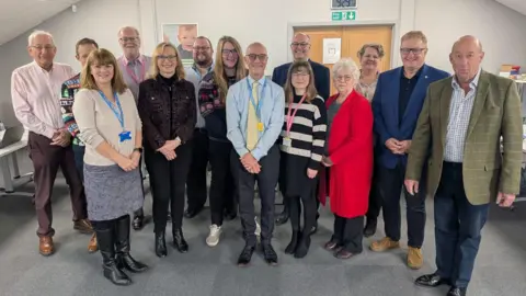 Hope House A large group of men and women are smiling at the camera. There are eight men and six women. Barbara is standing with them, with short white hair, glasses and a red jacket on