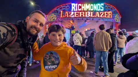 PT Vlogs Un hombre con un jersey camuflado extiende el brazo para tomarse una selfie con su hijo, un niño que sonríe y levanta el pulgar. Lleva un jersey amarillo que dice PT Vlogs y tiene otra selfie de él y su padre. Detrás de ellos hay gente parada alrededor de un letrero de neón que dice Richmonds Lazerblazer.