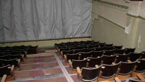 An interior shot of the original cinema inside. It shows a dark room with rows of dusty wooden chairs - four either side of the central steps - which leads down to the screen which is covered by a grey tarpaulin. 
