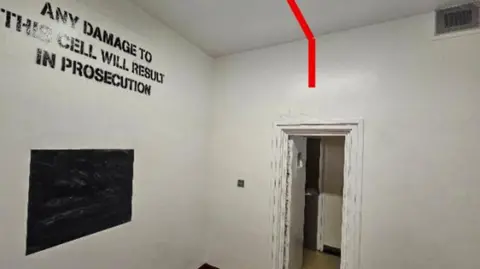 The interior of a holding cell with white-painted walls and a white ceiling. A doorway on the right-hand side leads into an adjoining corridor.
A thick red line has been digitally overlaid onto the image, running across the ceiling and down the wall above the doorway. Large black lettering painted onto the wall reads: ANY DAMAGE TO THIS CELL WILL RESULT IN PROSECUTION.