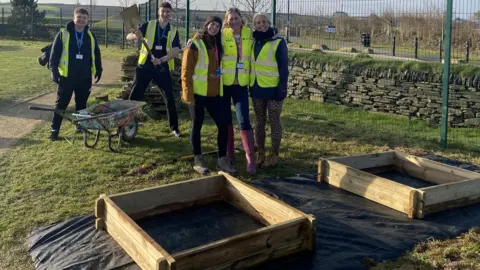 A group of three volunteers dressed in outdoor workwear and high vis jackets pose in front of the wooden frames of raised beds. Two other helpers stand behind with a wheelbarrow and a spade.