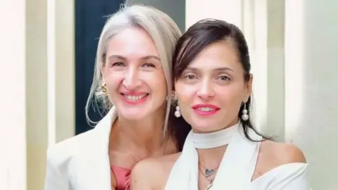 Tamuna Museridze Two smiling women standing close together. Nia (L) has long straight blonde hair that is tied back and is wearing a white jacket over a sleeveless, dusky pink dress; Tamuna (R) has long, dark, straight hair and is wearing a strapless white top with a white scarf.