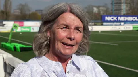 Siobhan is wearing a white shirt and has grey hair and is stood on the side of the rugby pitch. Signage with her husband's name on can be seen in the background as well as the green rugby pitch.