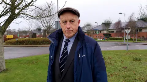 William is wearing a navy rain jacket, a blue shirt and a stripy tie. He is also wearing a flat cap. 