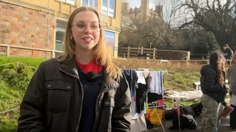 Livy Naylor looking slightly off camera while being interviewed. She has long light-coloured hair and is wearing large round-rimmed spectacles and a red top underneath a blue jumper and brown wax jacket.