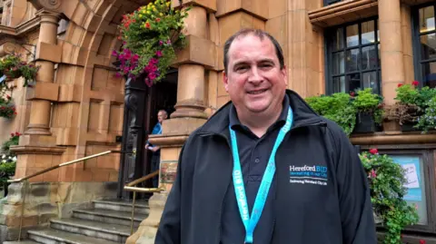 Local Democracy Reporting Service A man wearing dark clothing and a light blue lanyard is outside a building. He has dark hair and is looking at the camera. A number of steps are on the left of the image.