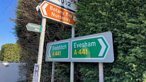 A series of road signs along the A441 Ridgway in Astwood Bank show the different destinations. Redditch is to the north, Evesham to the south. Smaller signs advertise the Monarch's Way footpath, as well as a local garden nursery. 