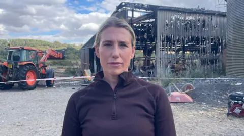 A woman looking at the camera. She has blonde hair which is tied back. She's wearing a brown jacket and is standing in front of a barn which has been burnt. There are holes in the sides and the front, and lots of ash on the floor. There is a red tractor in the background.