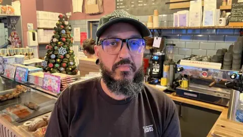 Phil Harrison/BBC A man in a black t-shirt and hat is standing inside a bakery.