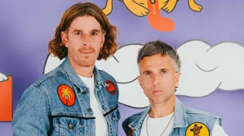 Two men in white T shirts and denim jackets with colourful badges stand in front of a purple background with white clouds on it