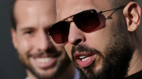 Octav Ganea/Reuters A head shot of Andrew Tate up close and just out of focus behind him is his brother Tristan. Sunshine is lighting their faces. Andrew is wearing sunglasses and has a black curly beard and Tristan has a similar beard. Andrew appears to be talking while Tristan is smiling.
