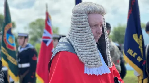 Deemster Graeme Cook wearing the red tunic of the deemsters and a long white wig. There are standard bearers behind him.