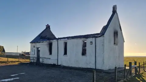 Firefighter in full protective gear and a bright yellow helmet spraying water onto the roof of a white building with severe fire damage. The roof is partially collapsed and charred, with smoke rising from the burnt section. A hose lies on the grass in front of the building, and the scene is set against an open field with a pale sunset sky