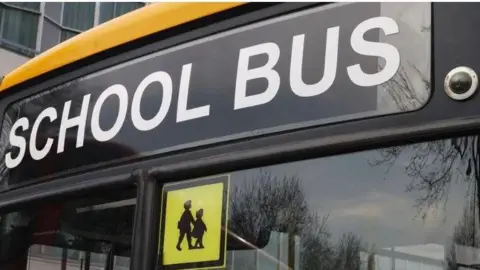 Getty Images Image shows the front windscreen of a school bus with the words school bus written at the top of the windscreen and in the middle of the windscreen at the top is a shadow picture of two children with a yellow background. 