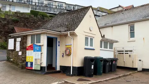 A toilet block in cream paint with a pointed roof and bins outside.