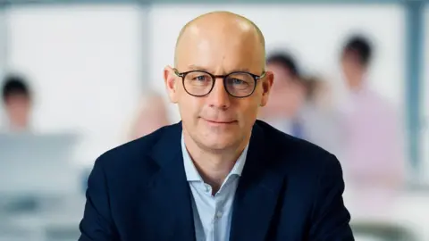 JT Group Daragh McDermott, JT Group's chief executive. He is a bald man wearing black-rimmed round glasses. He has a navy blazer and light blue stripey shirt with no tie.