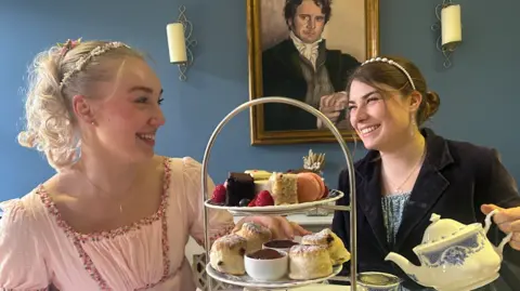 Jane Austen Centre Two ladies dressed in eighteenth century costume smiling at each other in a tea room with a large three-tiered plate of cakes and one of them pouring tea from a china pot. There is a painting of Colin Firth as Mr Darcy on the wall in the background