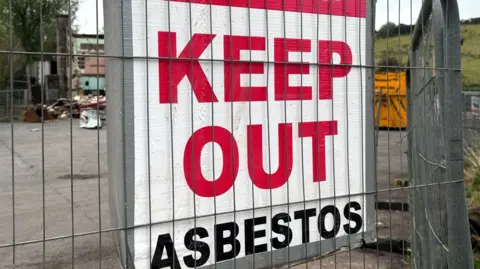 BBC A sign behind a wire fence. The sign has a white background - red text reads "KEEP OUT" and below it in black text "Asbestos"